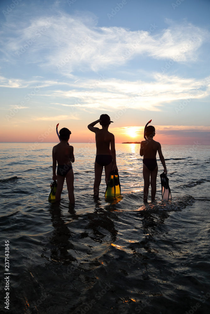 Silhouette-Rear View Of Three Boys Standing In The Sea With Diving Equipment And Looking At Horizon On Beautiful Sunset
