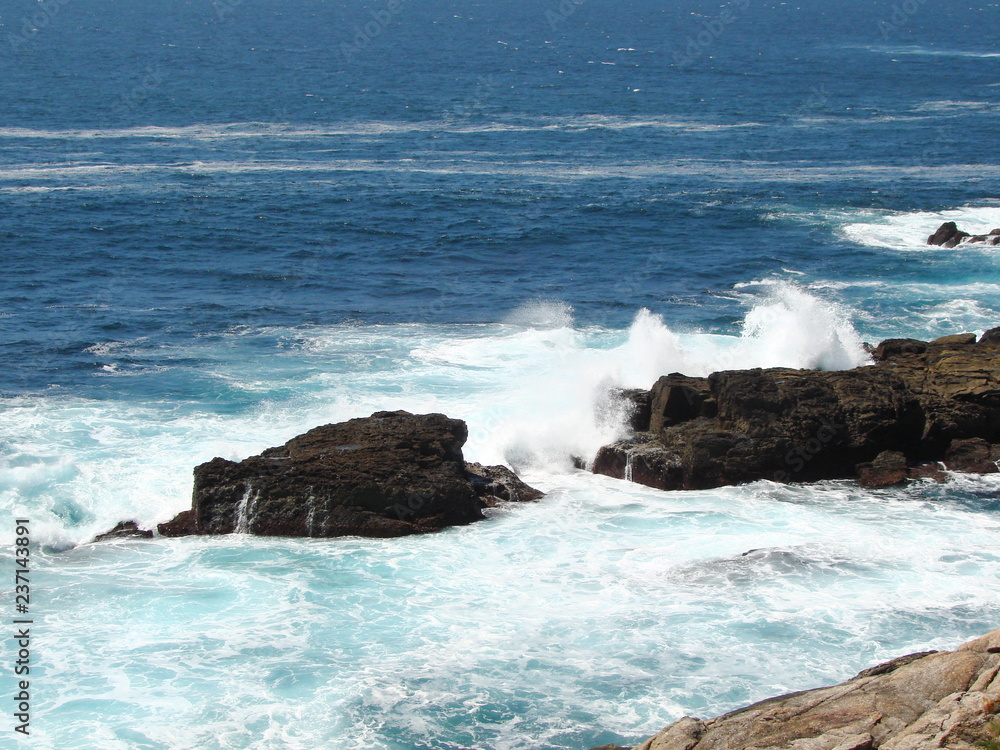 Panorama from the height of the coastal hill of white foam of ocean waves breaking on the rock against the background of dark-blue water surface.