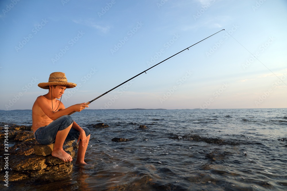 Boy Sitting On The Sea Rocks With A Fishing Rod And Patiently Waiting To  Catch A Fish Stock Photo
