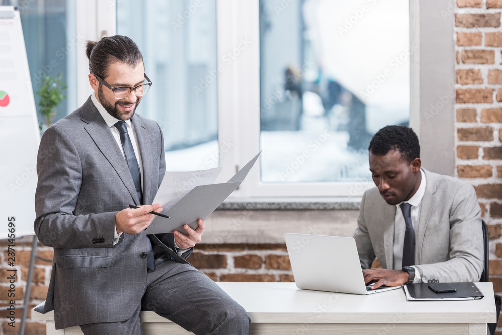 handsome businessman sitting on table and reading document while african american partner typing on laptop keyboard in office handsome businessman sitting on table and reading document while african american partner typing on laptop keyboard in office