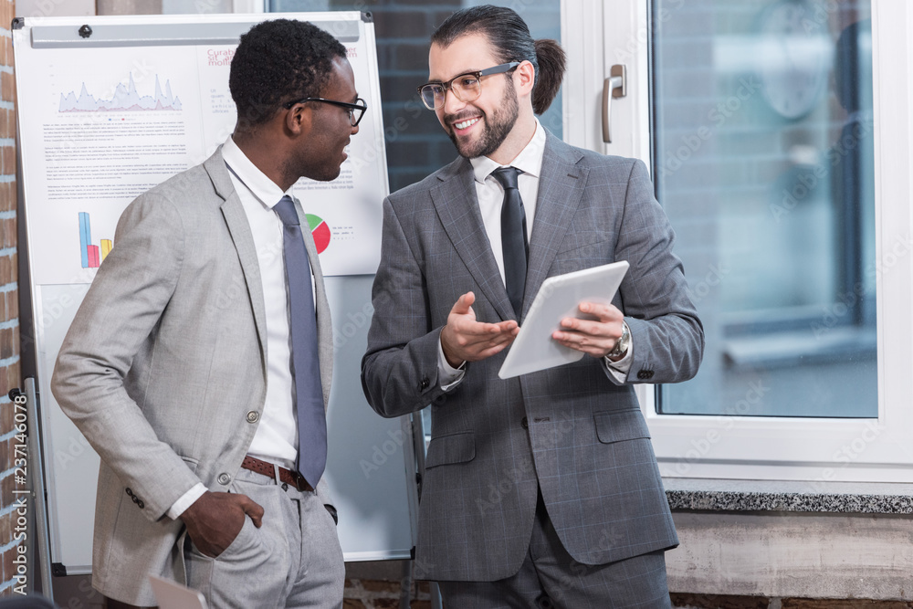 handsome businessman smiling and showing digital tablet to african american partner in office handsome businessman smiling and showing digital tablet to african american partner in office