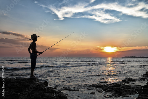 Young Boy Standing On The Rock And Fishing In The Sea At Beautiful Sunset 