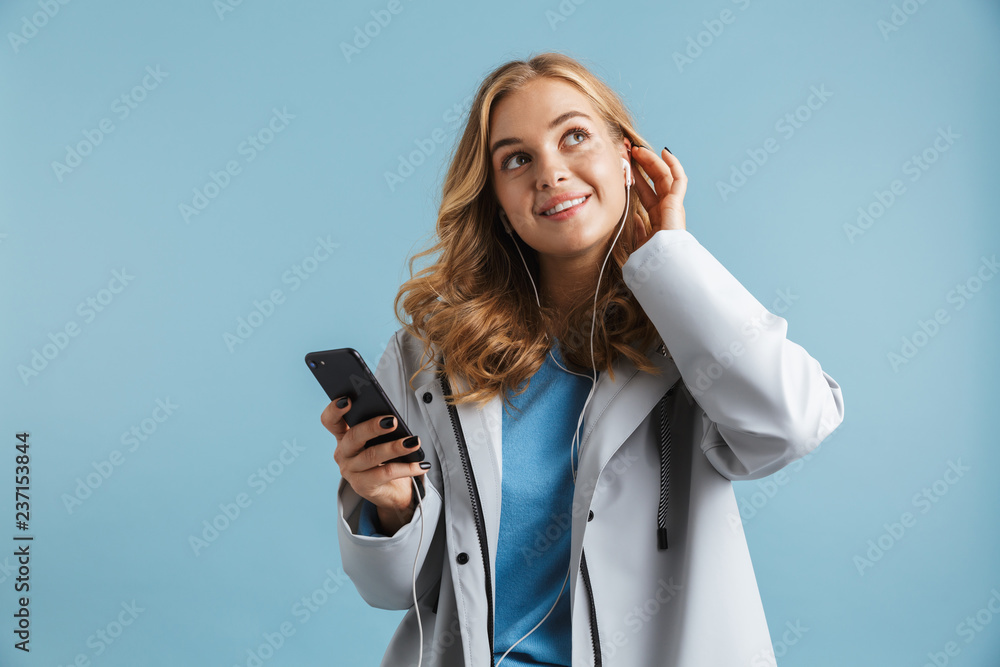 Image of young woman 20s wearing raincoat holding mobile phone and listening to music via earphones, isolated over blue background Image of young woman 20s wearing raincoat holding mobile phone and listening to music via earphones, isolated over blue background