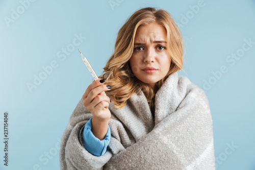 Image of ill upset woman 20s wrapped in blanket holding thermometer, isolated over blue background photo