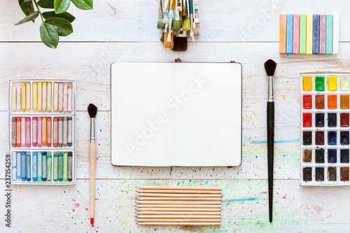 Creative work desk for self-expression, paint brushes, paintbox with watercolors, crayons, pencils and notebook paper on white wooden background, artistic workspace. Top view, flat lay, copy space