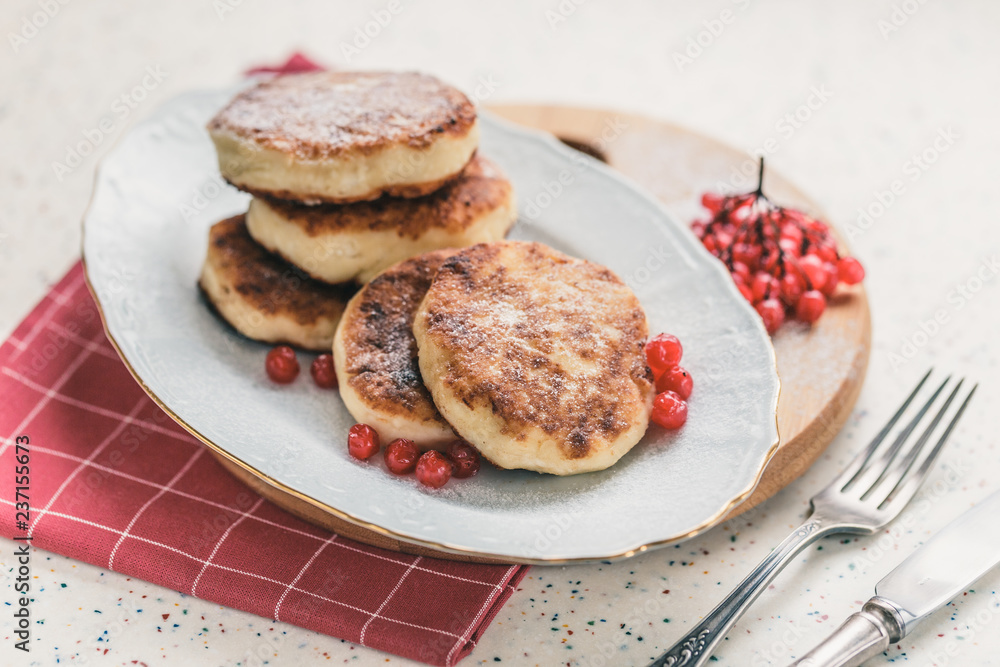 On a white table made of artificial stone is a plate with five cottage cheese pancakes and red berries . On the table is wooden cutting board and a red checkered napkin. Fork and knife. On a white table made of artificial stone is a plate with five cottage cheese pancakes and red berries . On the table is wooden cutting board and a red checkered napkin. Fork and knife.
