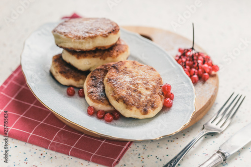 On a white table made of artificial stone is a plate with five cottage cheese pancakes and red berries . On the table is wooden cutting board and a red checkered napkin. Fork and knife.