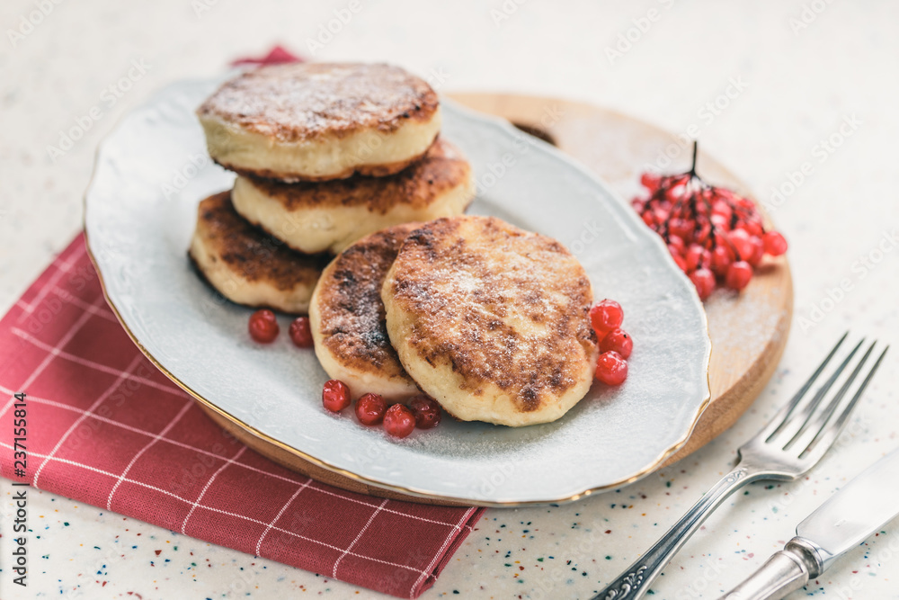 On a white table made of artificial stone is a plate with five cottage cheese pancakes and red berries . On the table is wooden cutting board and a red checkered napkin. Fork and knife.