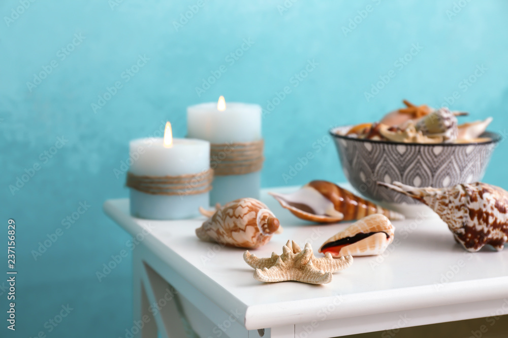 Different sea shells with burning candles on light table near color wall