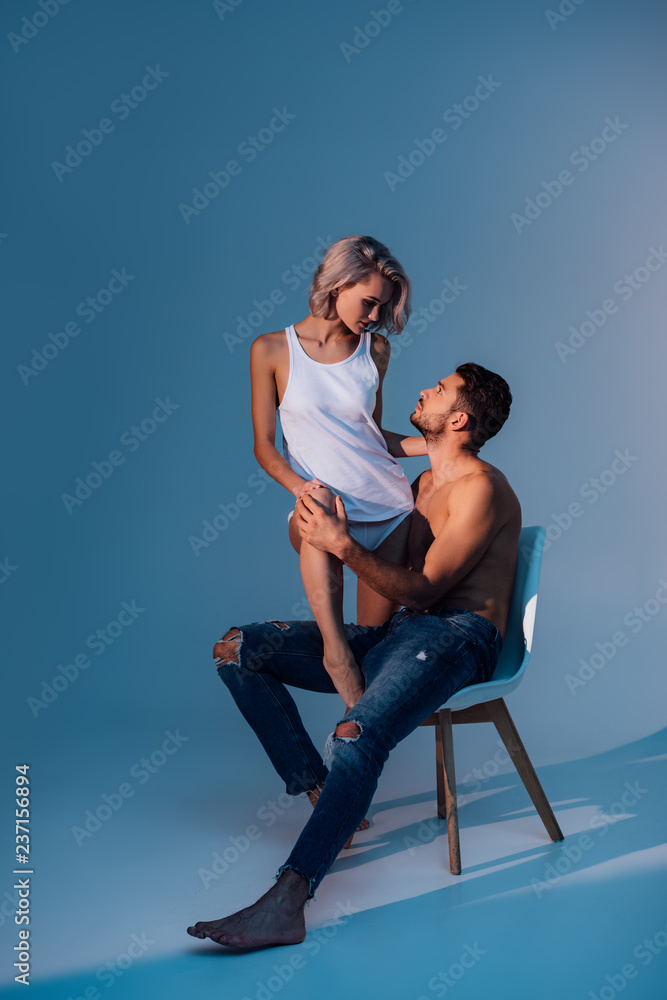shirtless man sitting on chair and looking at beautiful woman on dark blue background