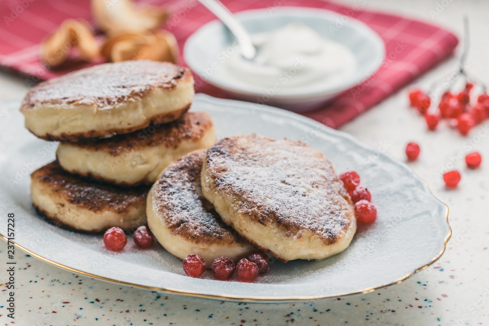 On a white table made of artificial stone is a plate with five cottage cheese pancakes and red berries . On the table is wooden cutting board and a red checkered napkin. Fork and knife. On a white table made of artificial stone is a plate with five cottage cheese pancakes and red berries . On the table is wooden cutting board and a red checkered napkin. Fork and knife.