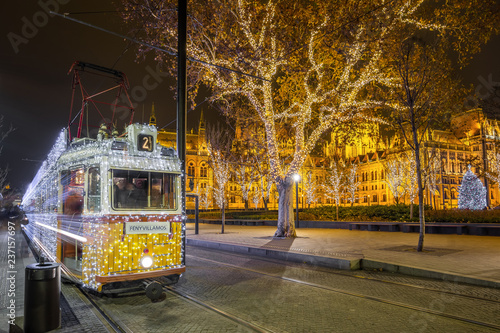 Festively decorated light tram (Fenyvillamos) on the move with Parliament of Hungary at Kossuth square by night. Christmas season in Budapest