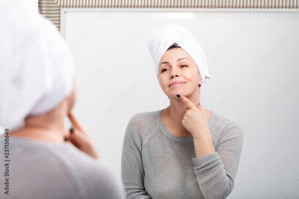Middle aged caucasian woman with bath towel on the head looking at reflection in mirror in the morning, admires herself and smiling Middle aged caucasian woman with bath towel on the head looking at reflection in mirror in the morning, admires herself and smiling