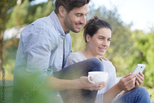  Relaxed young couple sitting together outside with mug and cellphone