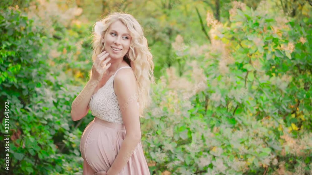 a pretty pregnant maid with blond wavy hair that winds fluttering around, in a bright summer morning forest, in a cute long delightful pink dress with a white lace top, runs her hand over her face a pretty pregnant maid with blond wavy hair that winds fluttering around, in a bright summer morning forest, in a cute long delightful pink dress with a white lace top, runs her hand over her face