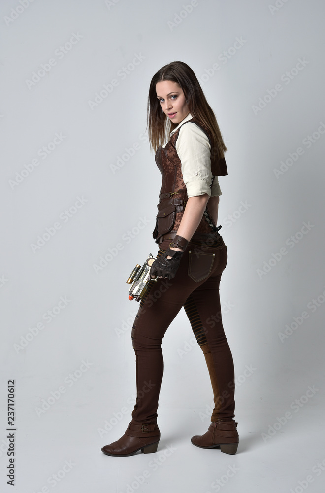 full length portrait of brunette girl wearing brown leather steampunk outfit. standing pose holding a gun on grey studio background. full length portrait of brunette girl wearing brown leather steampunk outfit. standing pose holding a gun on grey studio background.