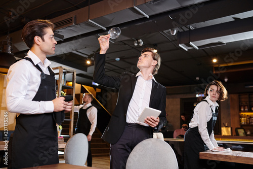 Serious professional restaurateur in black jacket holding tablet and checking cleanliness of wineglass while waiters wiping tables photo