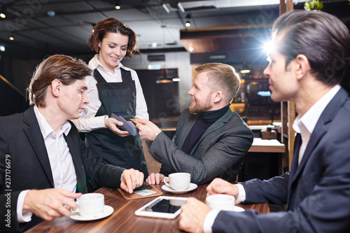 Positive attractive young waitress in black apron standing at table of guests and giving payment terminal to businessman who paying with contactless card in cafe photo