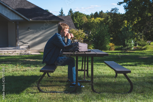 Upset teenager sitting at a picnic table in a park all alone. His backpack is lying on the table next to him.