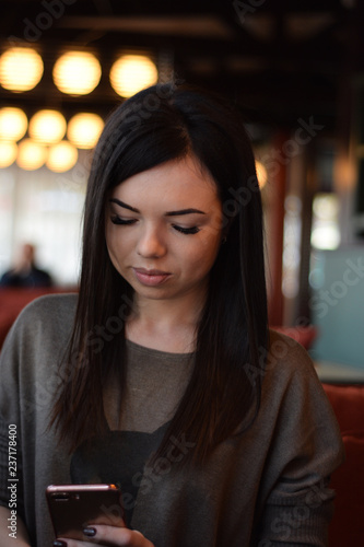 close-up portrait of a smiling beautiful girl with dimples on the cheeks in a cozy cafe against the window and bokeh. young woman is reading a message in the phone 