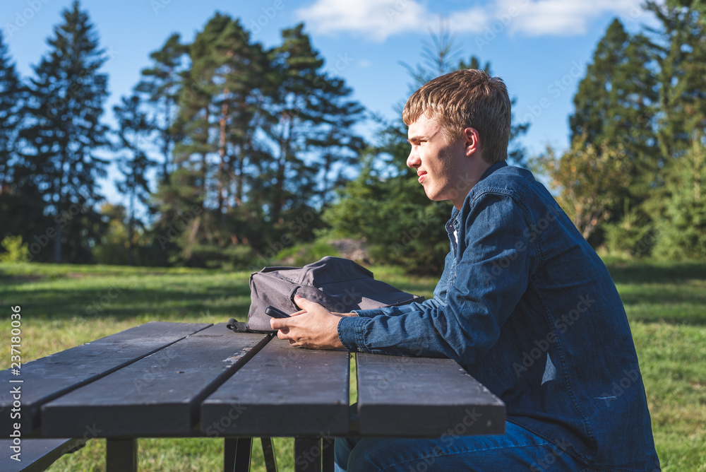 Fototapeta premium Concerned teenager sitting at a picnic table all alone with his phone.