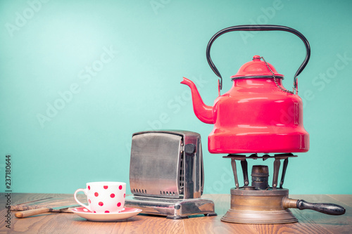 Retro classic red kettle on brass gas stove, a cup of tea, outdated bread toaster, kitchen board and vintage knife on oak wooden table in front mint green background. Old style filtered photo