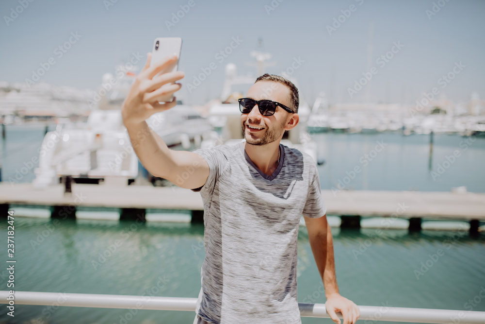 Young handsome man in sunglasses take selfie mobile phone on summer street with yacht harbor on background. Travel concept