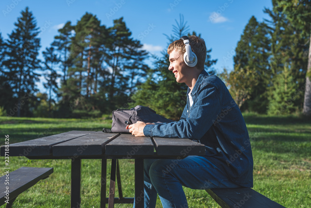 Fototapeta premium Happy teenager sitting at a picnic table in a park while listening to music.