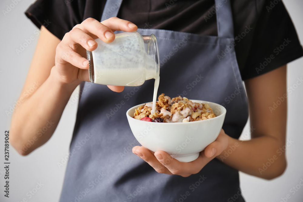 Woman pouring milk from jar into bowl with granola on light background, closeup