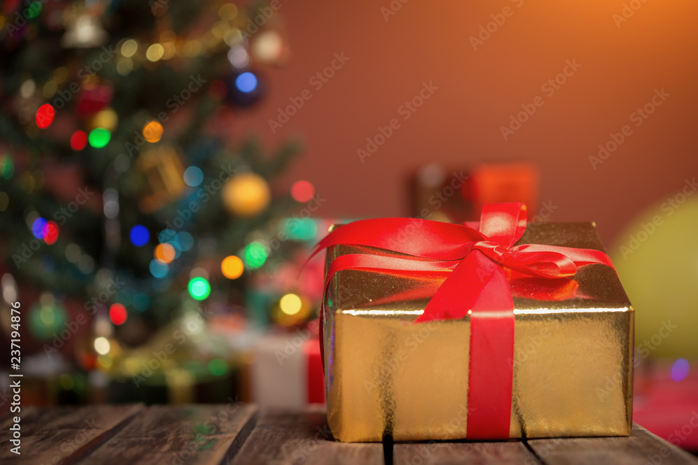 Beautiful golden gift box with red ribbon on wooden table at Christmas tree and lights background. Christmas concept
