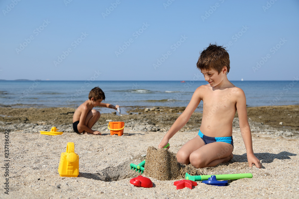 Twin Brothers Playing On The Beach, Making A Sand Castle With Plastic Toys For Sand Twin Brothers Playing On The Beach, Making A Sand Castle With Plastic Toys For Sand