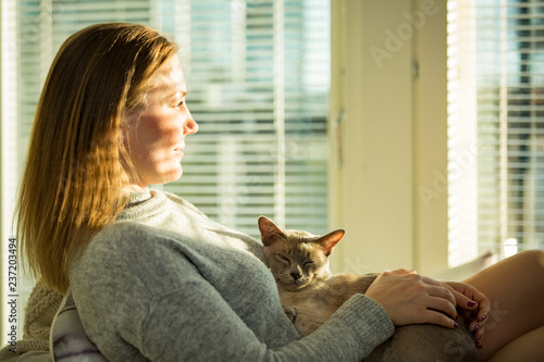 Woman sitting at home in a chair by the window with cup of hot coffee wearing knitted warm sweater, stroking cat on her laps. Cozy sunny room filled with light. beautiful morning behind big window.
