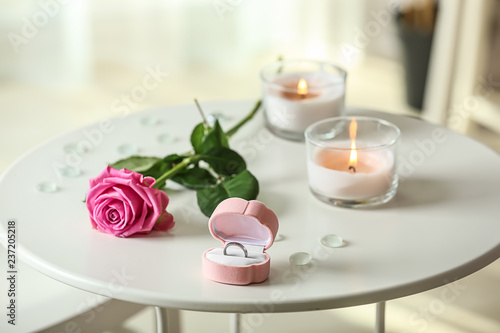 Engagement ring in box with rose and burning candles on white table