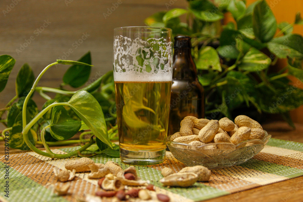 Beer in bottle and glasses and peanuts in crystal bowl. Table with vintage bamboo mat and liana vine in the background. Beer in bottle and glasses and peanuts in crystal bowl. Table with vintage bamboo mat and liana vine in the background.