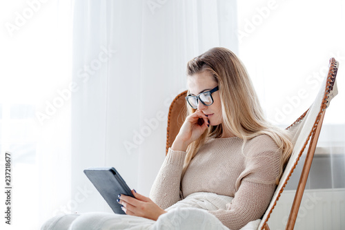 Young woman in glasses reading ebook using digital reader while sitting in comfortable armchair at home photo