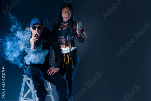 A man smokes an electronic cigarette. A man in the smoke. The guy and the girl smokes an electronic cigarette. Vape. Blacks fashion model. The girl photographs how a man smokes Vape. Vaping.