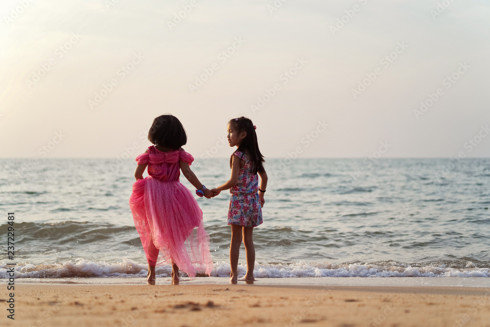 Two Asian kid girls holding hands each other on the beach while they are playing together. Seen from their back in the evening sunset. Vacation and holiday on beach concept.