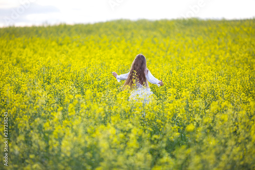 A girl in a white dress runs along a flowering rapeseed field on a sunny day. © Valiantsina