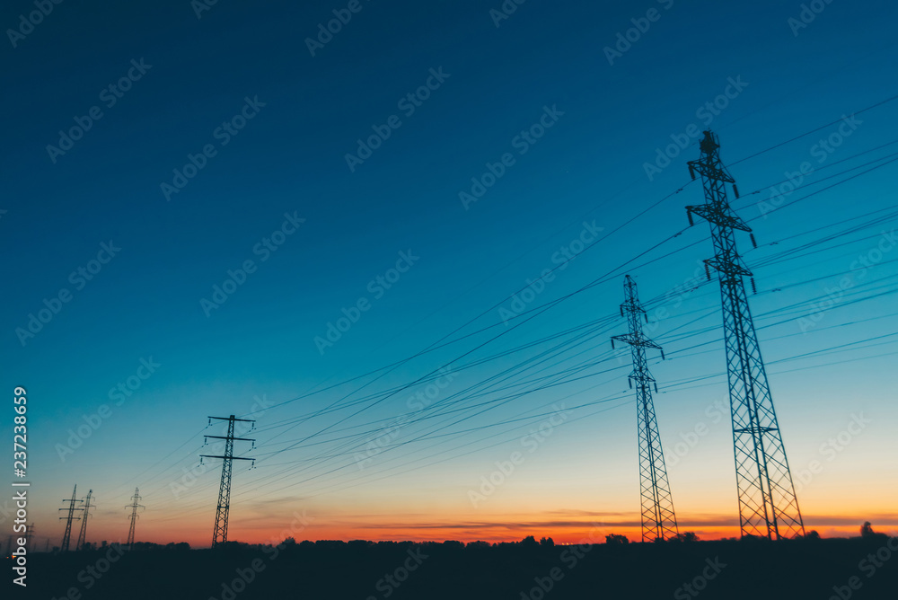 Power lines in field on sunrise background. Silhouettes of poles with wires at dawn. Cables of high voltage on warm orange blue sky. Power industry at sunset. Many cables in picturesque vivid sky.
