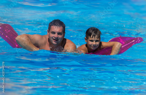 Father and his teen son relaxing at the weekend, swimming in a pool on an inflatable pool raft. © Kate