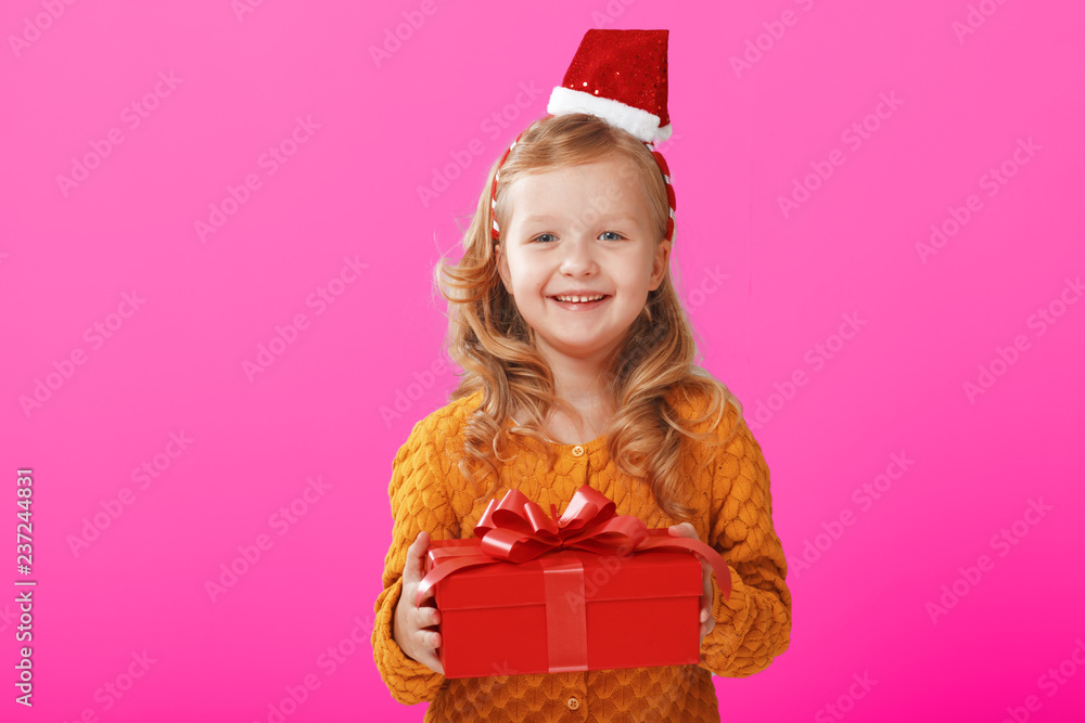 Happy little child girl in a warm knitted sweater and cap Santa Claus is holding a red box with a gift on a pink background. The concept of celebration, giving and receiving a gift Happy little child girl in a warm knitted sweater and cap Santa Claus is holding a red box with a gift on a pink background. The concept of celebration, giving and receiving a gift