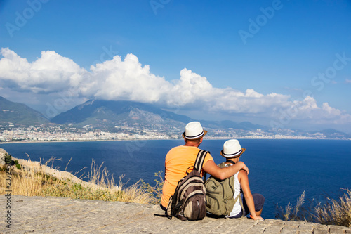 Young father and son with backpacks are sitting on the beach against the backdrop of the sea. Family travel concept