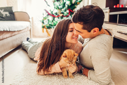 Couple in love lying by Christmas tree and playing with cat at home. Man and woman kissing