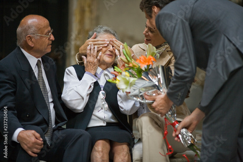 Mid-adult woman covering her senior mother's eyes as her brother presents a bouquet of flowers while sitting near her senior father. photo