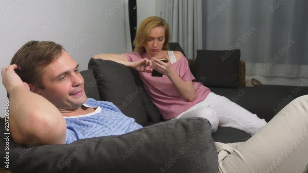 Couple crisis. Man and woman spend the evening in the living room on the couch. the woman uses the phone, the man is upset that she does not pay attention to him. Couple crisis. Man and woman spend the evening in the living room on the couch. the woman uses the phone, the man is upset that she does not pay attention to him.