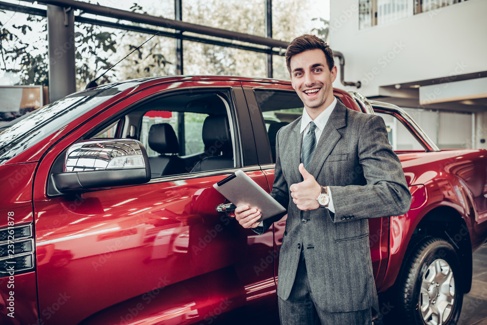 Handsome smiling salesman in formal wear with tablet in hand looking on camera and shows thumb up on new car background