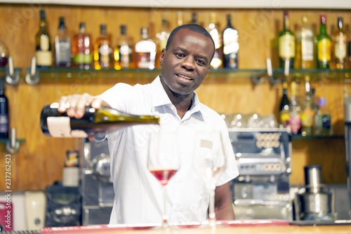 African barman men standing  pours red wine into a glass from a bottle. Shelves with bottles of alcohol in the background. Focus on the bartender.