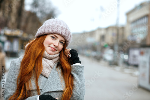 Closeup shot of wonderful ginger woman with long hair wearing knitted cap and scarf walking at the city. Empty space