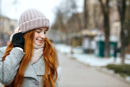 Closeup shot of lovely ginger woman with long hair wearing knitted cap and scarf walking at the city. Empty space
