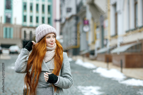 Urban portrait of pretty ginger girl with long hair wearing warm clothes walking at the city. Empty space
